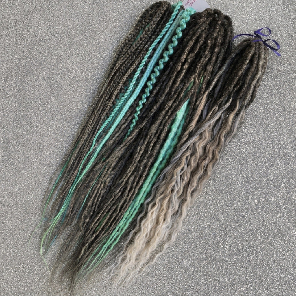 Three bundles of synthetic dreadlocks or box braids laid flat on a textured gray background. One bundle is dark brown with bright turquoise accents, one is dark brown with turquoise and light blue accents, and one is dark brown with blonde and gray ombré ends.